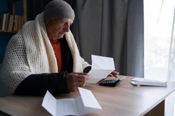 Older man wrapped up in a blanket as well as wearing a hat and gloves to keep warm inside his house, he's reading his fuel bill.